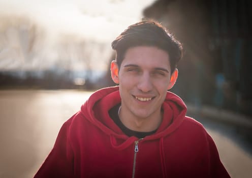 A young man wearing a red hoodie smiles directly at the camera in this portrait photo. His expression is cheerful and inviting, exuding a sense of friendliness and approachability.