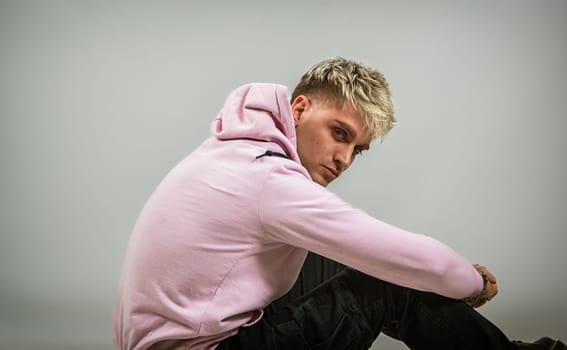 A man wearing a pink hoodie is sitting on the ground in this photo. He appears relaxed and lost in thought, with his legs crossed and hands resting on his knees.