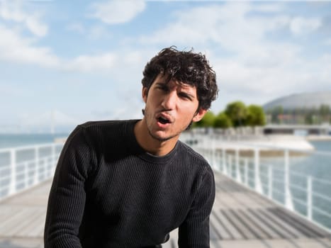 A man standing on a pier, looking surprised or hurting from pain, with his mouth open and eyebrows raised. The background shows water and boats.