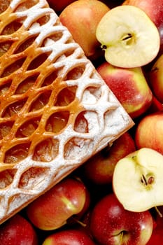 Closeup of classic apple galette with lattice top dusted with icing sugar, presented alongside whole and sliced fresh ripe apples on checkered tablecloth