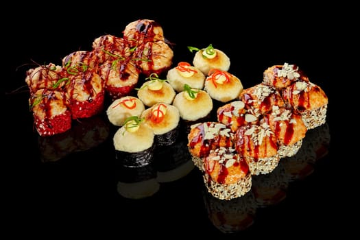 Set of baked sushi rolls - uramaki in sesame and norimaki topped with creamy cheese hats drizzled with spicy mayo and unagi sauce, garnished wish parmesan crumbs and red chili threads on black surface
