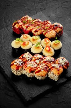 Set of appetizing baked sushi rolls topped with creamy cheese hats seasoned with spicy mayo and unagi sauce, garnished with parmesan crumbs, vegetable shavings and red chili threads on slate board