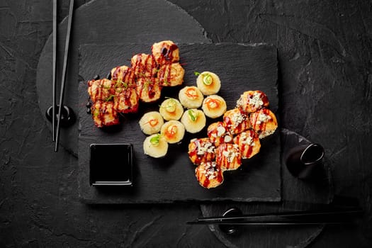 Top view of appetizing baked sushi rolls topped with cheese hats seasoned with spicy mayo and unagi sauce, garnished with parmesan crumbs and red chili threads served on slate plates with soy sauce