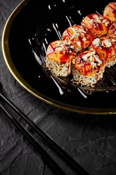 Exquisite baked uramaki sushi rolls in sesame seasoned with tangy unagi sauce sprinkled with Parmesan crumbs served on black and gold platter accompanied with chopsticks against dark stone surface