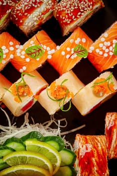 Top view of vibrant assorted sushi rolls with salmon, eel, tuna, mamenori garnished with masago, sesame and airy rice balls accompanied by shredded daikon, cucumber and lime slices on black background