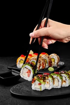 Female hand with chopsticks taking from slate board appetizing sushi roll filled with cream cheese, shrimp and tomato topped with hiyashi wakame, red tobiko and unagi sauce against black background