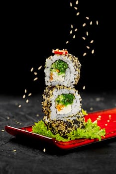 Stack of light vegetarian sushi rolls with cream cheese, carrots, hiyashi wakame, cucumber and chili pepper sprinkled by sesame seeds served on fresh lettuce leaf on vibrant red plate against black background