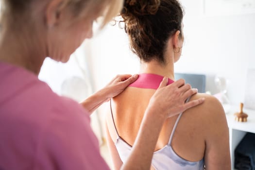 Back view of crop unrecognizable careful female therapist applying kinesiology tape on neck of female patient during physiological therapy in modern clinic