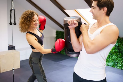 Young smiling female fighter in boxing gloves punching heavy bag during intense training with personal instructor in sports center