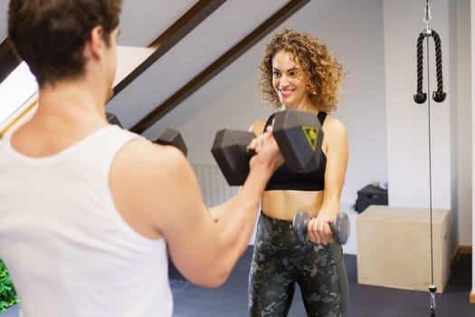 Young motivated female athlete lifting heavy dumbbells during workout with male partner in gym with professional equipment