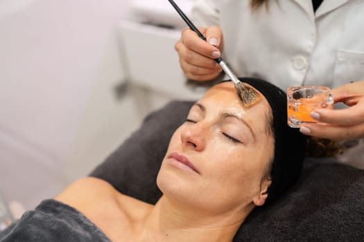 High angle of anonymous beautician with brush applying moisturizing treatment on face of relaxed woman from container in beauty salon