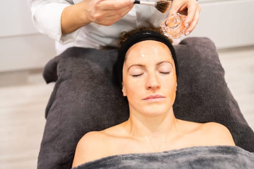 High angle of lady relaxing on bed while anonymous cosmetologist applying moisturizing product with brush from container in beauty center