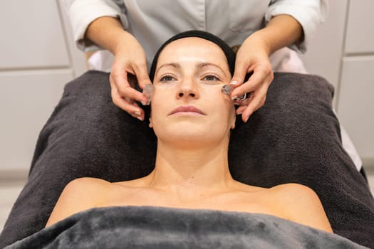 High angle of crop anonymous cosmetician in robe smoothing skin of woman with natural crystals during anti aging procedure in beauty salon