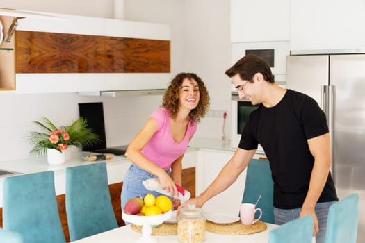 Positive adult couple in casual clothes, while standing near dining table smiling female looking at camera pouring juice and looking down male in eyeglasses holding glass during conversation