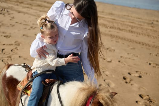 Caucasian blonde little child girl feeling worried and hugging her mother while riding pony on the sandy beach. Mon and daughter enjoying happy moments together