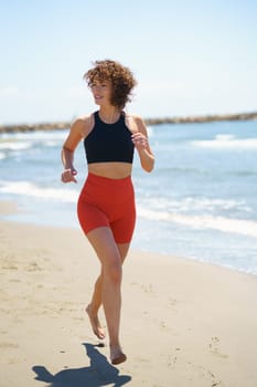 Full body of smiling barefoot young female in sportswear, with curly hair jogging on sandy beach and looking away while warming up near waving sea in sunny summer day