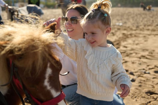 Young mother and her little kid girl, a lovely daughter smiling and stroking the pony. Mother and daughter admiring together a pony horse. Family engaging in outdoors activities with little child