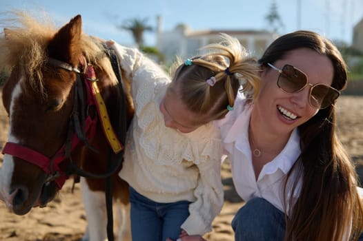 Happy young mother and daughter admiring together a pony horse. Young family engaging in outdoors activities with little child. People. Leisure and hobbies. Active healthy lifestyle