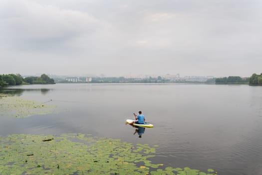 Male kayaker among water lilies on a calm lake, the cityscape faintly visible against the foggy background