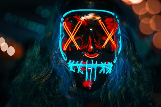 Man with long disheveled hair in a lighting neon glowing mask. Halloween dress-up party. Anonymous person is a cyber criminal.