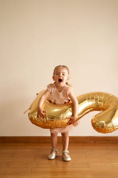 Little laughing girl with an inflatable number two in front of her stands against the wall. High quality photo