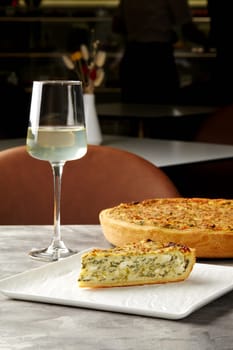 Slice of piquant cheese and spinach quiche with crispy shortbread crust served with glass of white wine in cozy cafe. Elegant dining setting