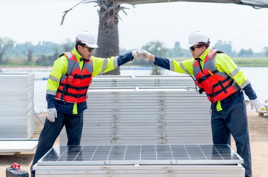Two Asian technician worker stand beside solar cell panel and show hands bump to each other in concept of green energy system and success of work.