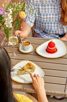 Female friends enjoying light snacking with savory tart and sweet red dessert with wine and coffee at outdoor cafe table, cropped image