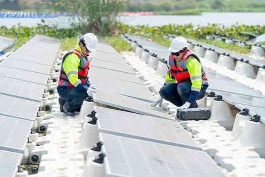 Two professional technician workers sit on footpath of row of solar cell panels and check or maintenance in area of green energy power plant workplace over water reservoir.
