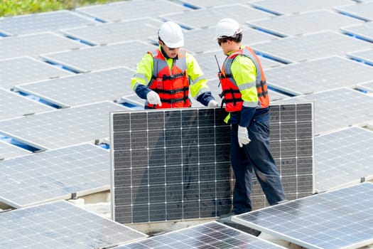 Two Asian professional technician workers with safety uniform stand and help to check or maintenance solar cell panels in area of workplace over water reservoir.