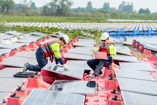 One professional technician worker use laptop to work with other co-worker touch to check and maintenance solar cell panel in workplace over water reservoir.