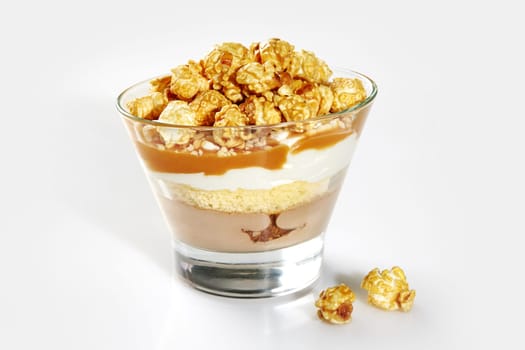 Tempting sweet layered trifle with sponge cake, panna cotta, rich caramel sauce, whipped cream, and caramel coated popcorn in clear dessert glass on white background