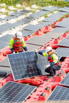 Vertical image of two technician workers help to set up or install solar cell panel into base over water reservoir.