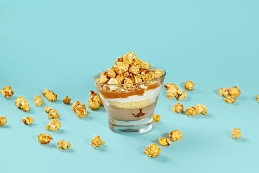 Tempting layered dessert with sponge cake, caramel sauce and whipped cream topped with golden caramelized popcorn served in glass on blue background