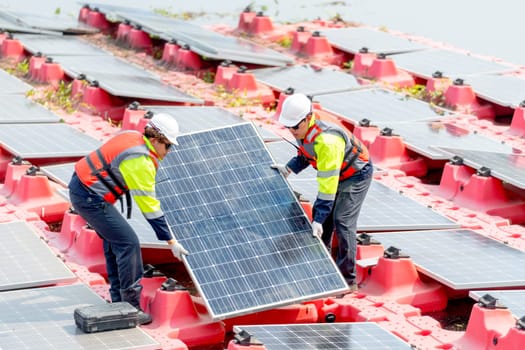 Two professional technician workers hold and carry solar cell panel to set up or install to the base over area of water reservoir.