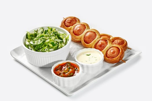 Tray of freshly baked pigs in blanket, green side salad, and assorted dips served for breakfast on white background