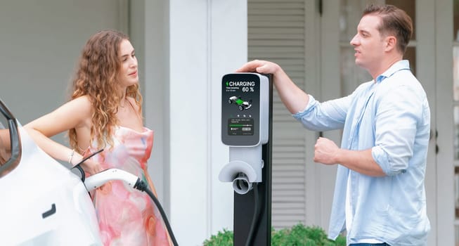 Happy and lovely couple with eco-friendly conscious recharging electric vehicle from EV home charging station. EV car technology utilized for residential home to future sustainability. Synchronos