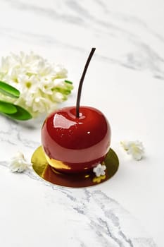 Tempting cherry-shaped pastry topped with red glossy glaze decorated with white flowers presented on gold cardboard cake base against marble backdrop. Collections of signature handmade desserts