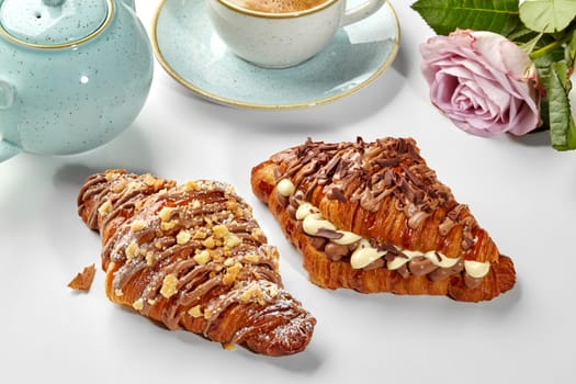 Two flaky golden croissants with custard cream filling topped with chocolate icing, flakes and caramelized crumbles, accompanied with cup of coffee and delicate rose on white background