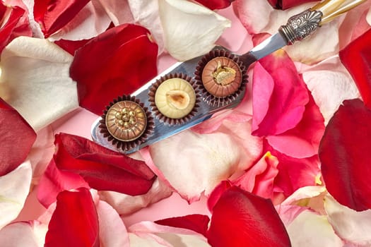 Three gourmet chocolates decorated with hazelnuts, caramel crumbs and golden pearls resting on ornate silver serving spoon, surrounded by vibrant red and soft pink rose petals