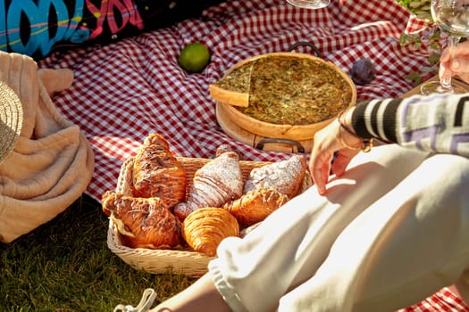 Cozy summer picnic scene on sunny day on green grass with sweet fluffy croissants in wicker basket, homemade savory quiche and fresh fruits scattered on checkered blanket, cropped shot