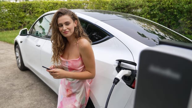 Young woman use smartphone to pay for electricity at public EV car charging station in nature. Modern environmental and sustainable automobile transportation lifestyle with EV vehicle. Synchronos