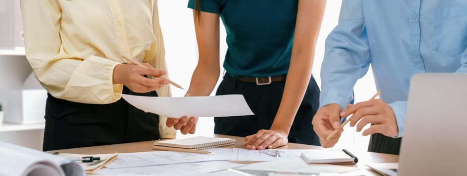 Architect team discuss about blueprint design at modern meeting room while her coworker working on laptop on table with blueprint and architectural equipment scattered around. Closeup. Delineation.