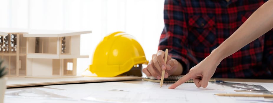 Cropped image of cooperative architect team decide and work together on meeting table with house model, safety helmet and architectural plan scatter around. Closeup. Focus on hand. Burgeoning.