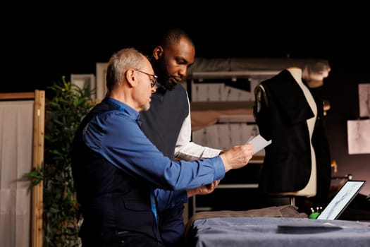 Master menswear collection creator and apprentice working on sartorial suit adjustments. Tailors modifying fashion sketches, trying different clothing combinations on mannequin in atelier