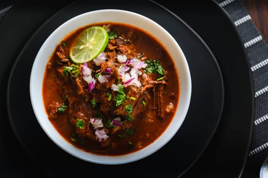 White bowl filled with delicious red Mexican beef birria stew on black plates. Served with red onion, cilantro and lime. Traditionally from Jalisco and central Mexican states.