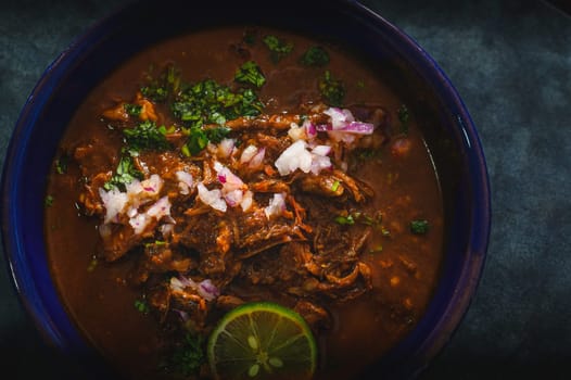 Blue bowl filled with delicious red Mexican beef birria stew on blue background. Served with red onion, cilantro and lime. Traditionally from Jalisco and central Mexican states.