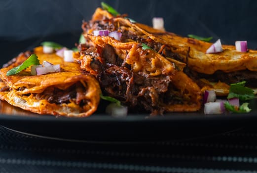 3 crispy, juicy beef birria tacos stacked on a black plate. Appetizing layout of amazing Mexican food in Tijuana, Mexico.