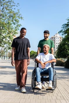 Vertical portrait of young disabled Black man in a wheelchair and his male friends walking together in the city. Friendship concept.
