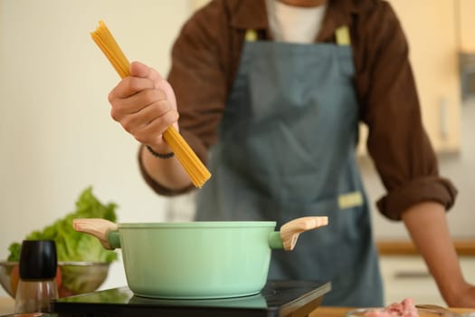 Young man in apron putting raw spaghetti pasta into pot with boiling water.